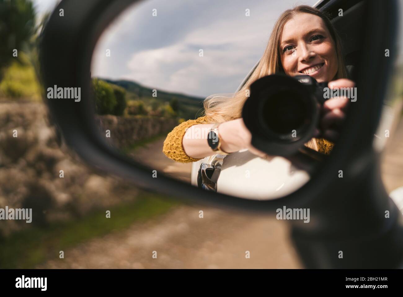 Spiegelung im Spiegel der lächelnden jungen Frau mit Kamera, die aus dem Autofenster gelehnt ist Stockfoto