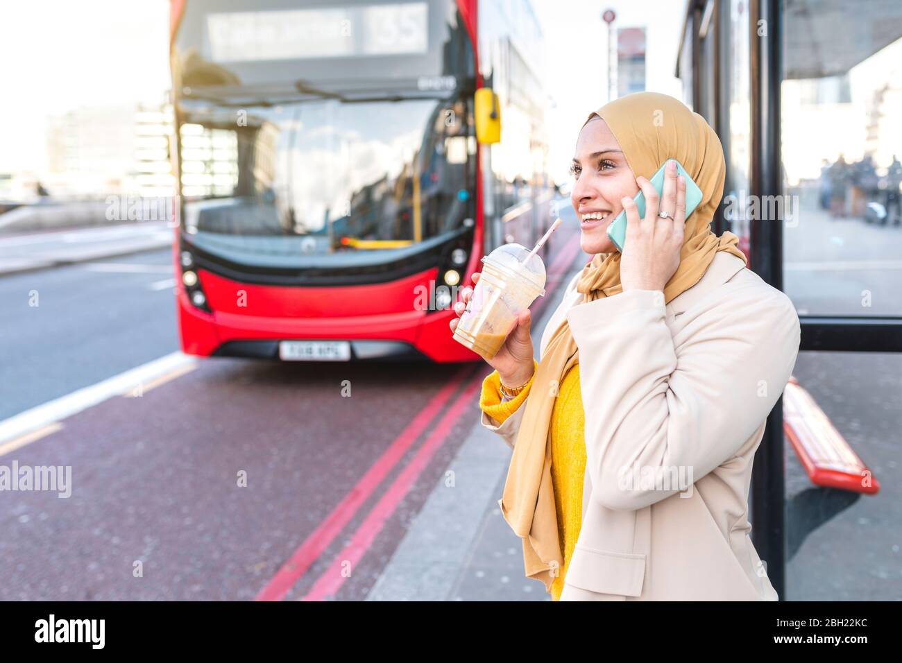 Porträt einer lächelnden jungen Frau am Telefon an der Bushaltestelle warten Stockfoto
