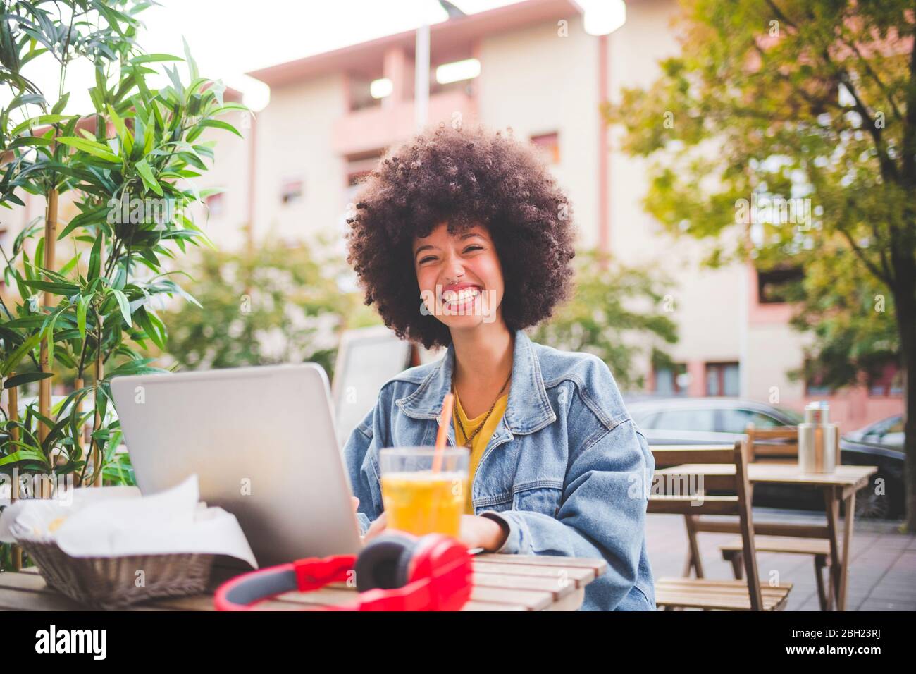 Glückliche junge Frau mit Afro-Frisur mit Laptop in einem Café im Freien in der Stadt Stockfoto