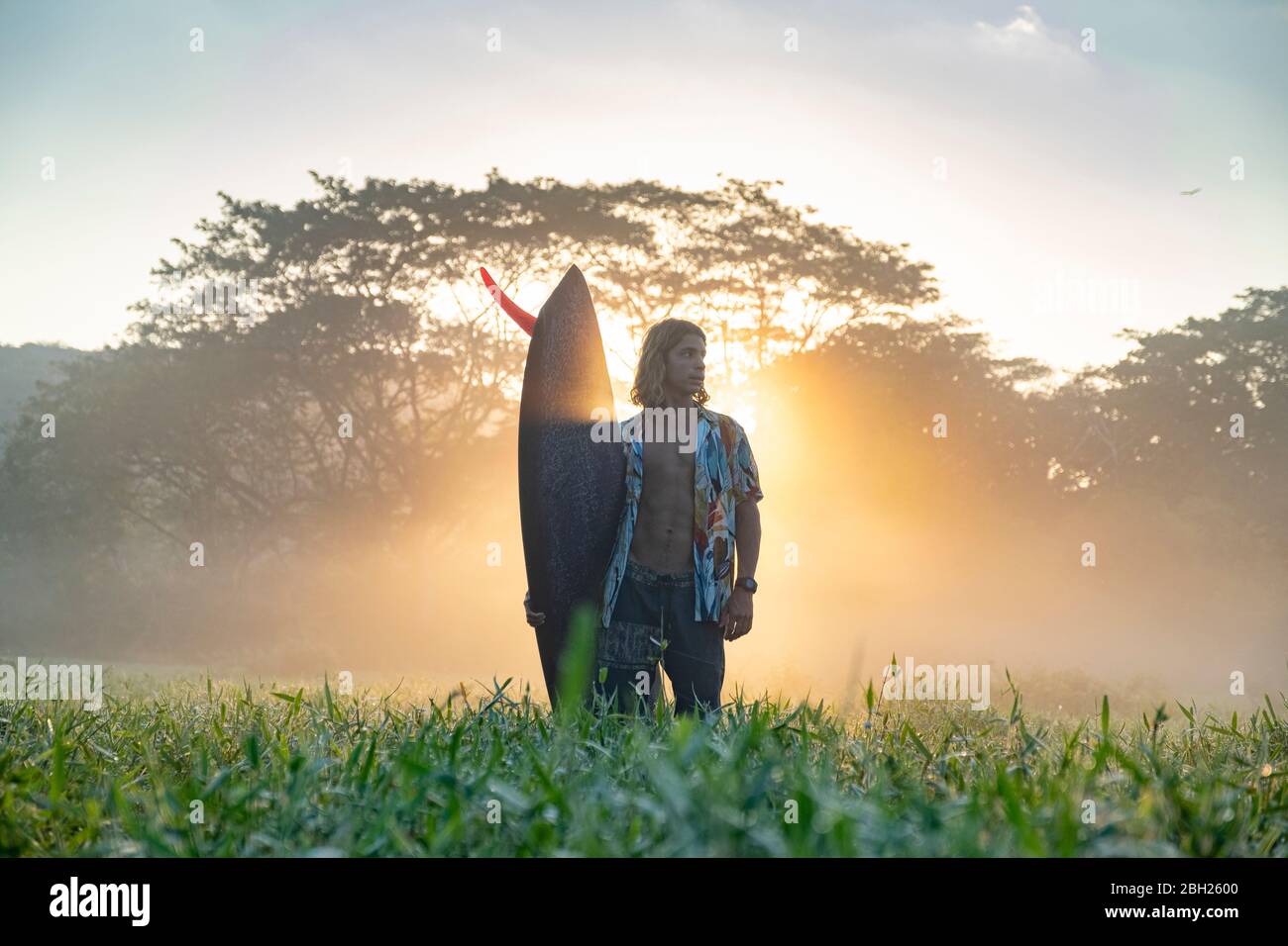 Junger Mann mit Surfbrett, der bei Sonnenaufgang auf einer Wiese steht, Costa Rica Stockfoto