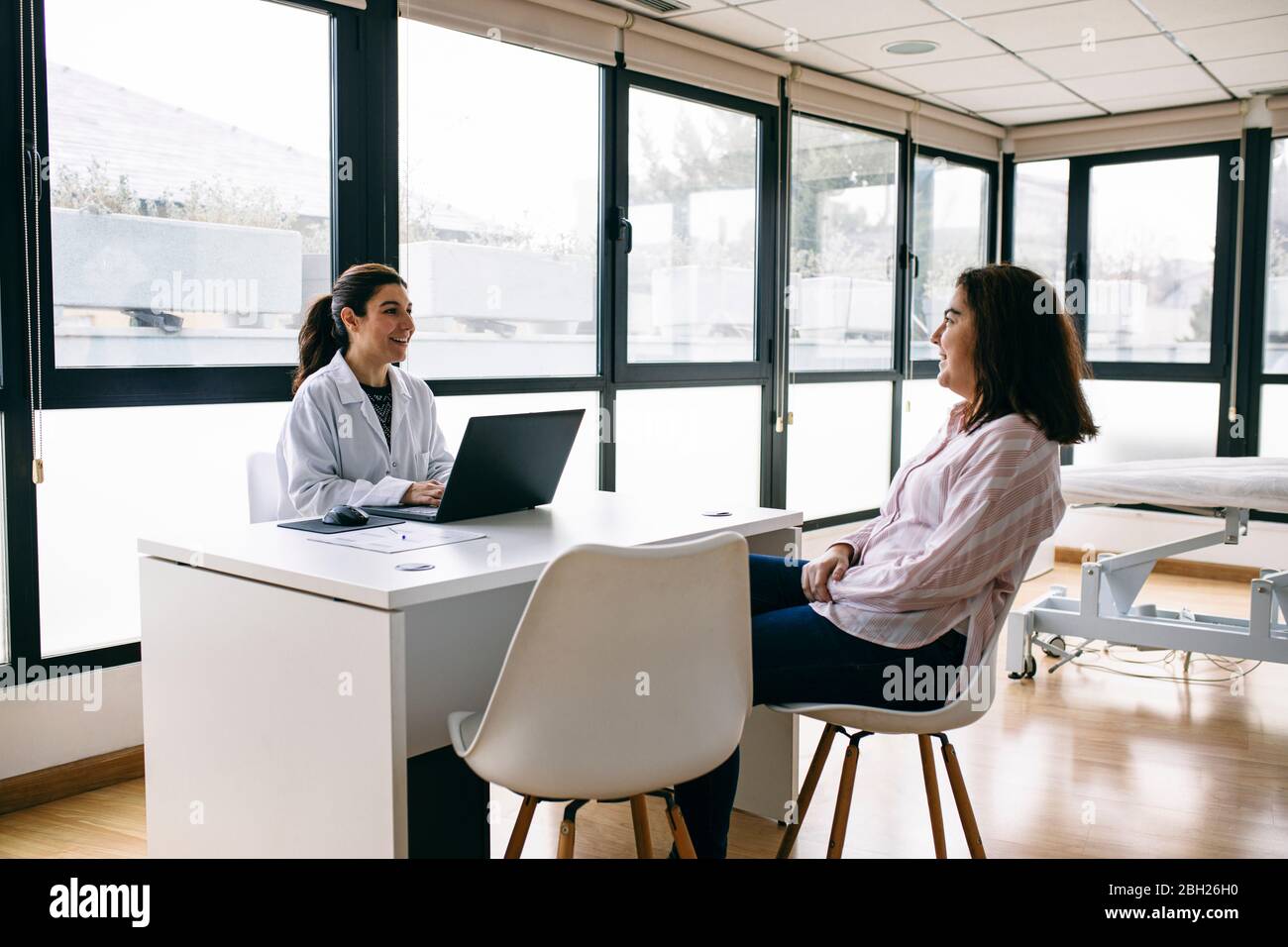 Lächelnder Arzt mit Laptop im Gespräch mit dem Patienten in der medizinischen Praxis Stockfoto