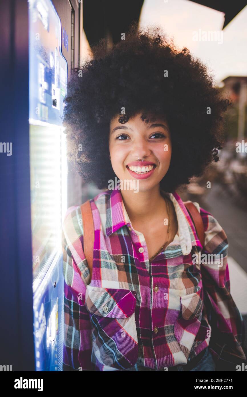 Porträt einer glücklichen jungen Frau mit afro Frisur lehnt sich an Automaten in der Stadt in der Dämmerung Stockfoto