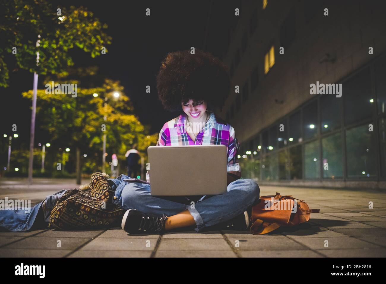 Junge Frau mit Afro-Frisur sitzt auf dem Boden mit Laptop in der Stadt in der Nacht Stockfoto