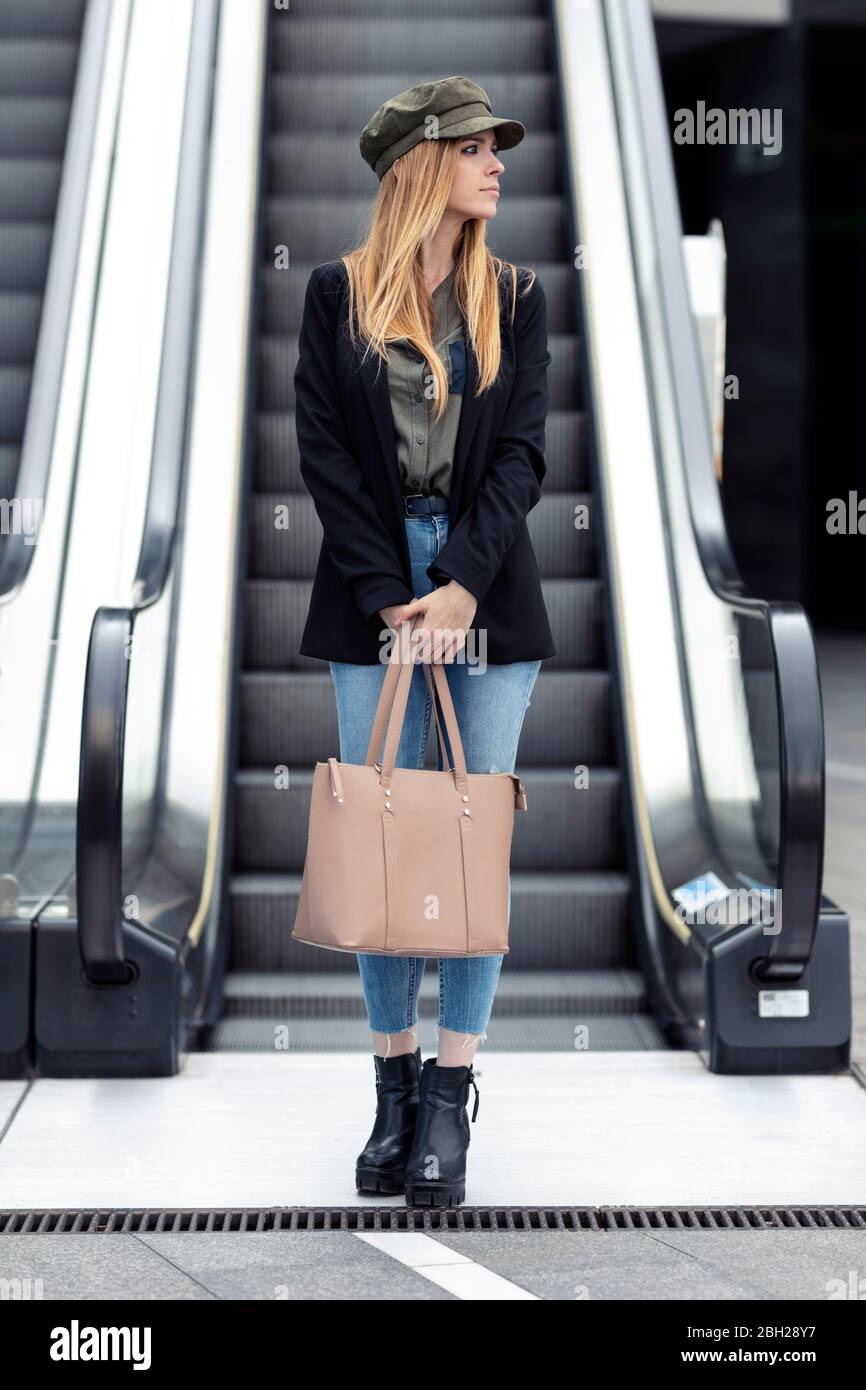 Portrait einer blonden jungen Frau mit Tasche, die vor einer Rolltreppe steht Stockfoto