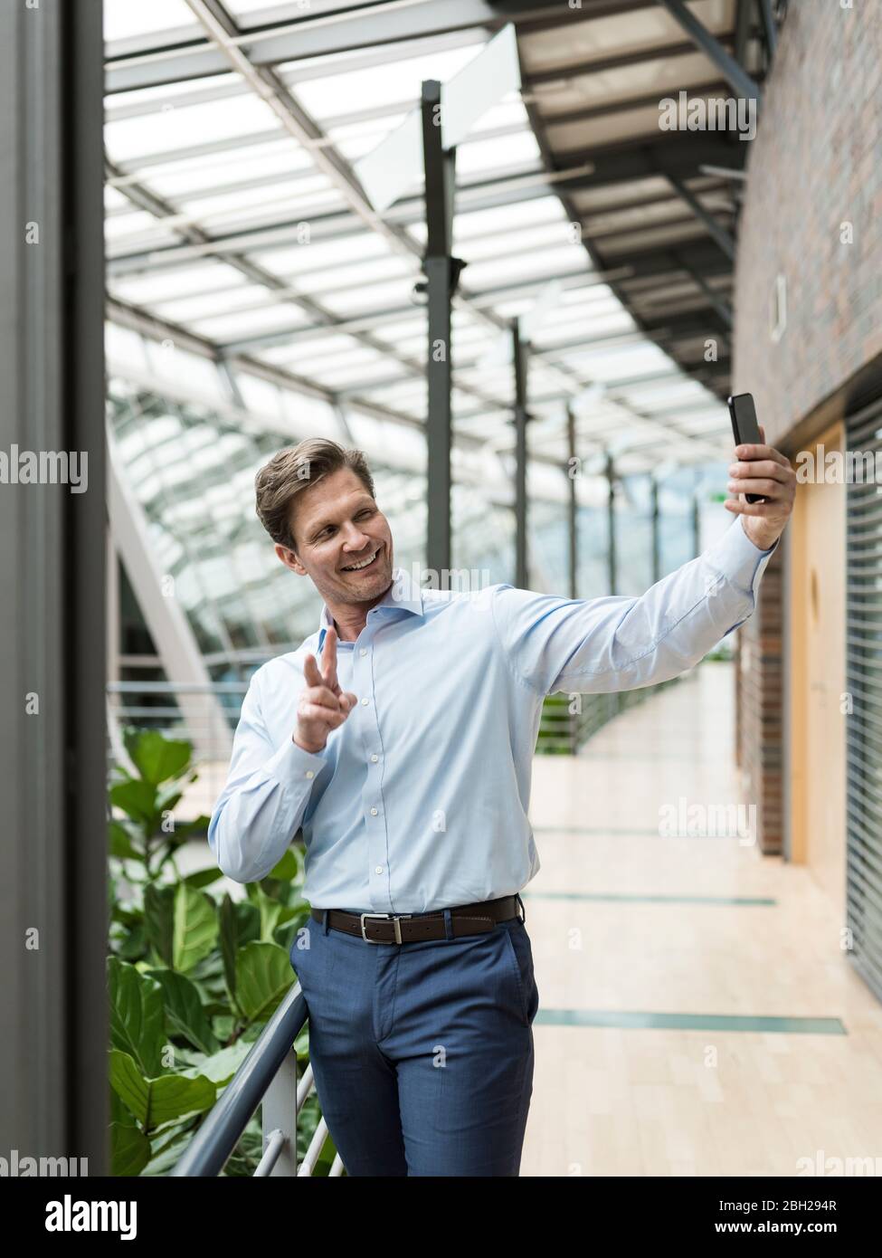 Geschäftsmann im grünen Atrium, Smartphone Selfie, macht Siegeszeichen Stockfoto