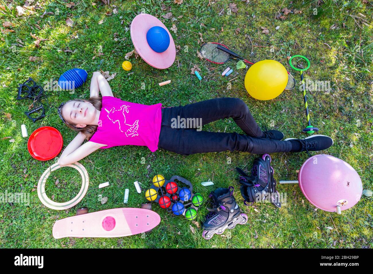 Mädchen liegt auf Gras, mit geschlossenen Augen, umgeben von Spielgeräten Stockfoto