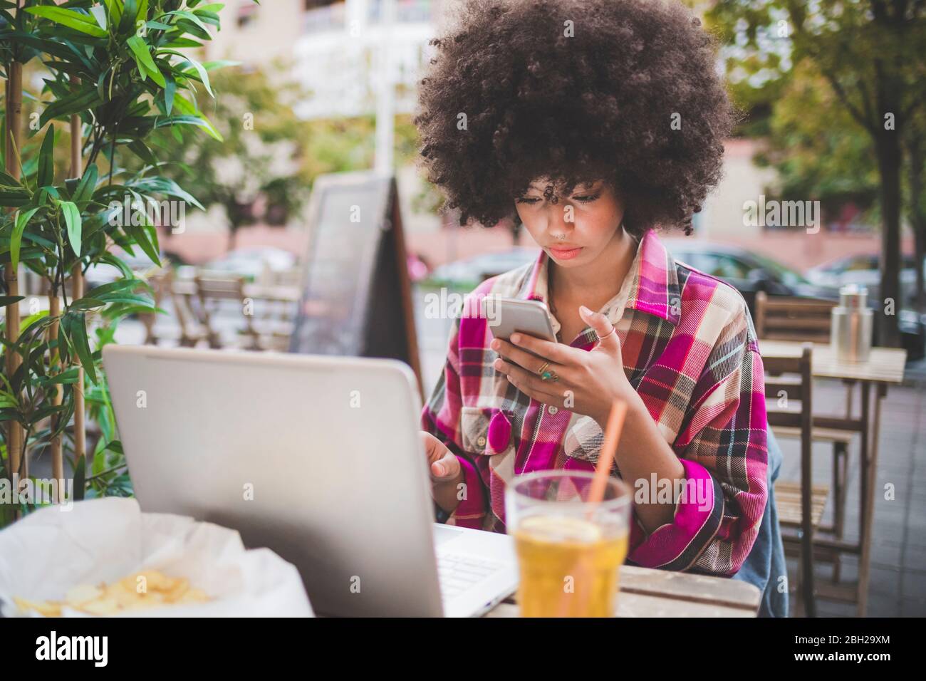Junge Frau mit Afro-Frisur mit Smartphone und Laptop in einem Café im Freien in der Stadt Stockfoto