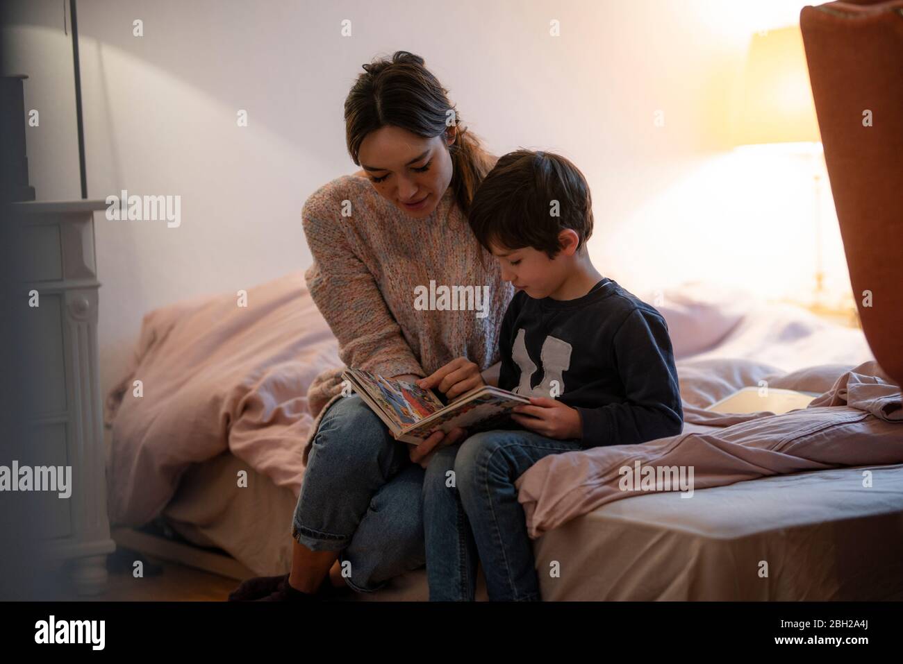 Mutter und kleiner Sohn sitzen zusammen auf dem Bett und schauen sich Bilderbuch an Stockfoto