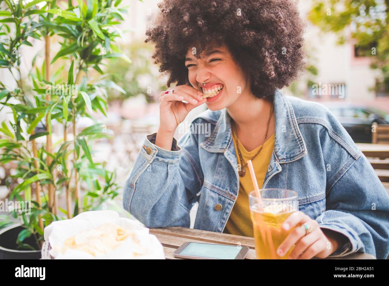 Glückliche junge Frau mit Afro-Frisur, die Chips in einem Café im Freien in der Stadt isst Stockfoto
