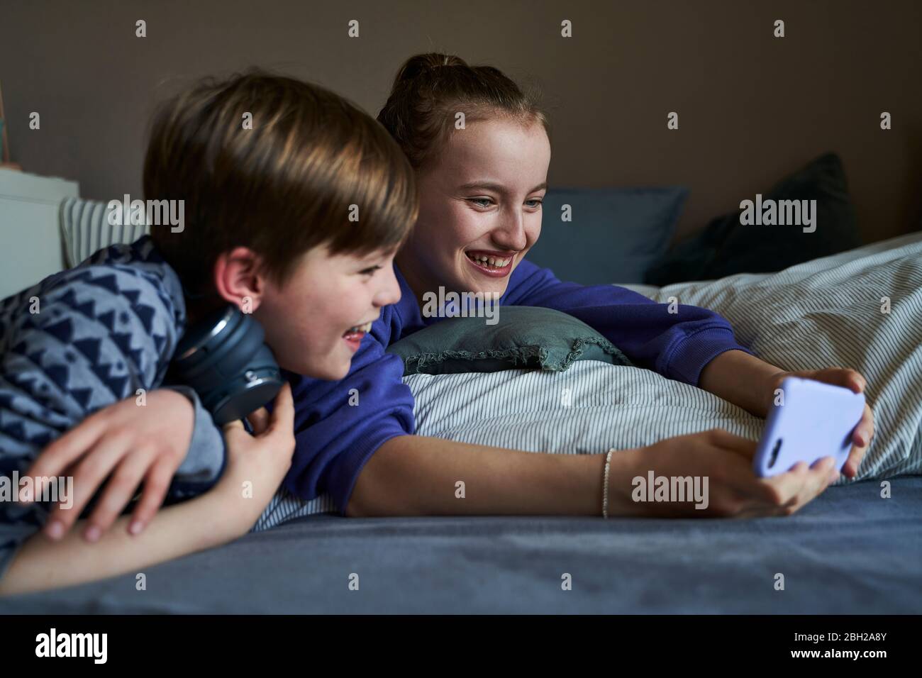 Lachende Sibblings liegen zusammen auf dem Bett mit Smartphone für Video-Chat Stockfoto