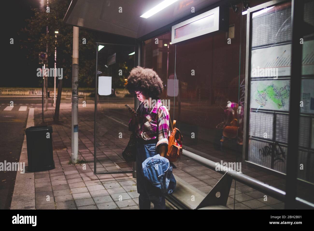 Junge Frau mit Afro-Frisur mit Smartphone an der Bushaltestelle in der Stadt Stockfoto