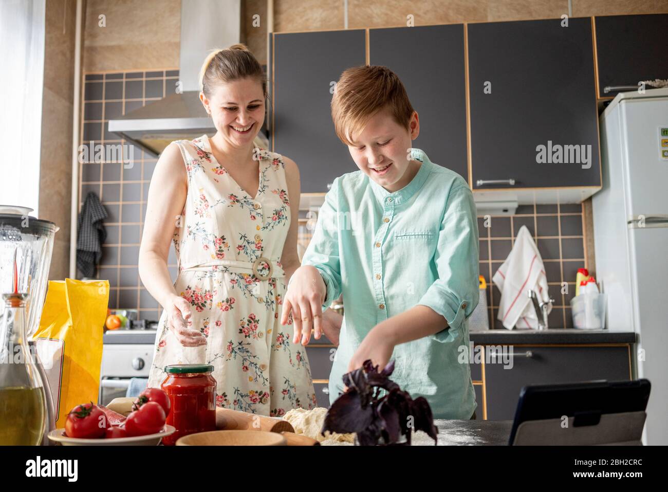 Lächelnder Junge kniet Teig in der Küche, während seine Mutter zuschaut Stockfoto