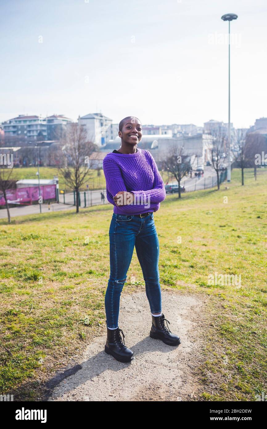 Selbstbewusste Frau steht im Park, lächelnd, mit gekreuzten Armen Stockfoto