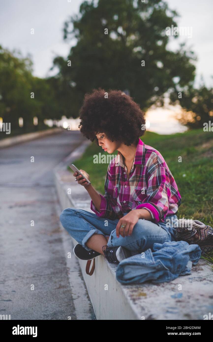 Junge Frau mit Afro-Frisur sitzt auf Bordstein mit Smartphone Stockfoto