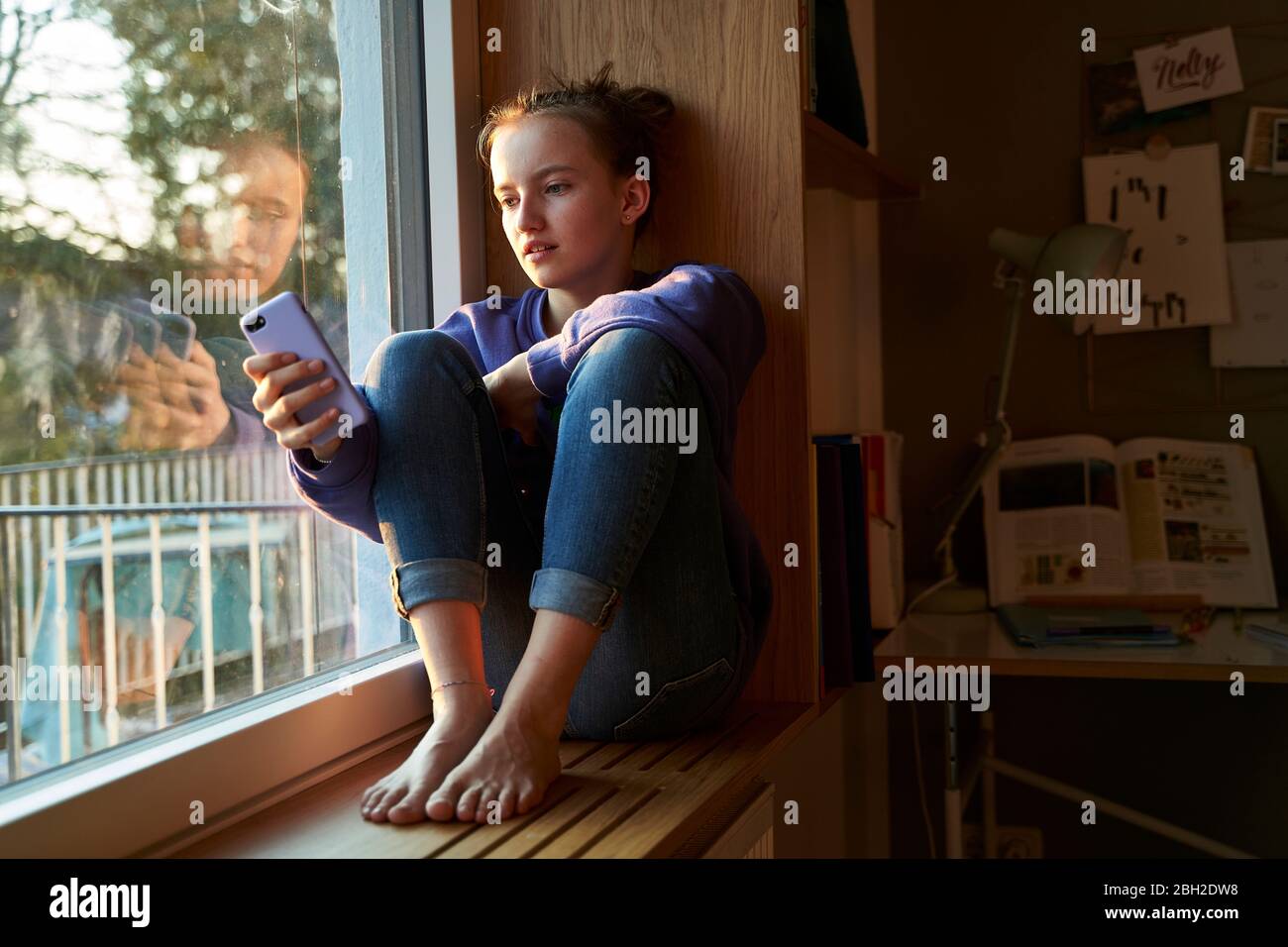 Mädchen, das barfuß auf der Fensterbank am Abend sitzt und Smartphone anschaut Stockfoto