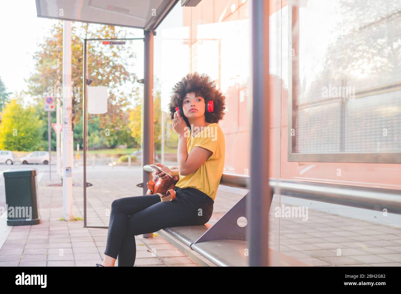 Junge Frau mit Afro-Frisur Musik mit Kopfhörern an der Bushaltestelle in der Stadt hören Stockfoto