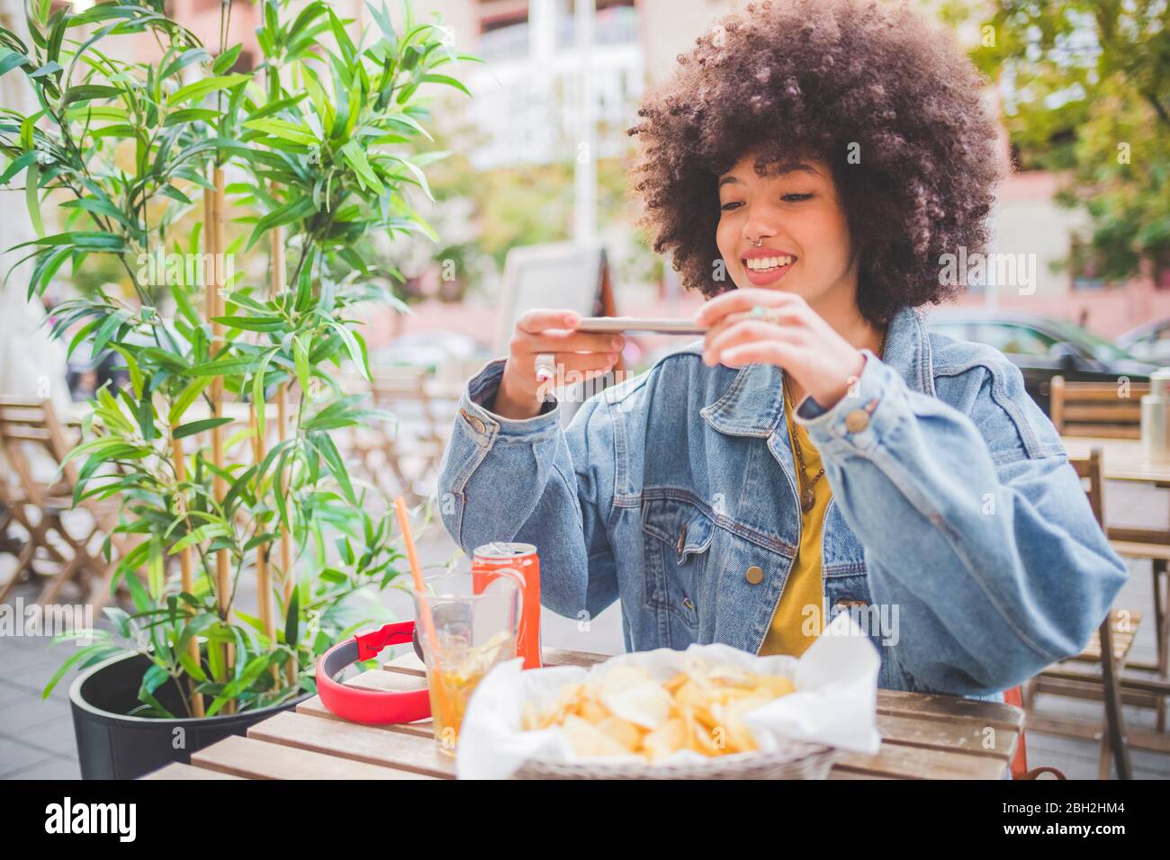 Lächelnde junge Frau mit Afro-Frisur, die Smartphone-Fotos in einem Café im Freien in der Stadt macht Stockfoto