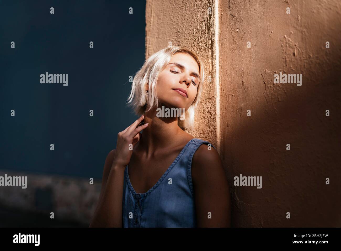 Portrait einer jungen blonden Frau, die sich an der Wand lehnt und das Sonnenlicht genießt Stockfoto