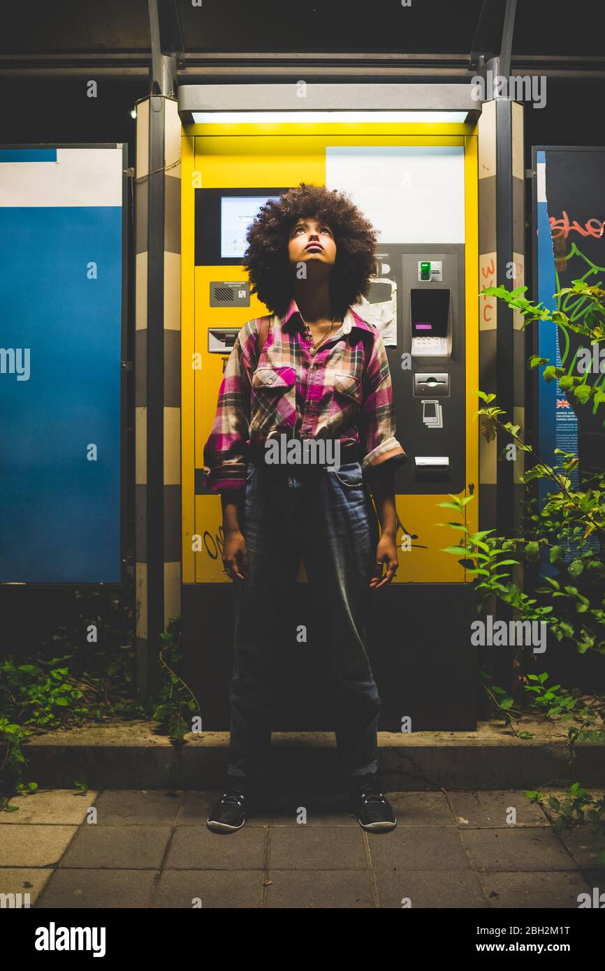 Junge Frau mit Afro-Frisur, die am Ticketautomaten in der Nacht aufschaut Stockfoto