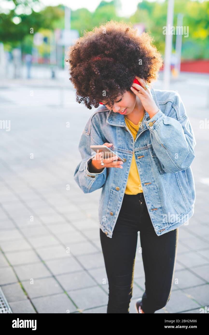 Junge Frau mit Afro-Frisur mit Smartphone und Kopfhörer in der Stadt Stockfoto