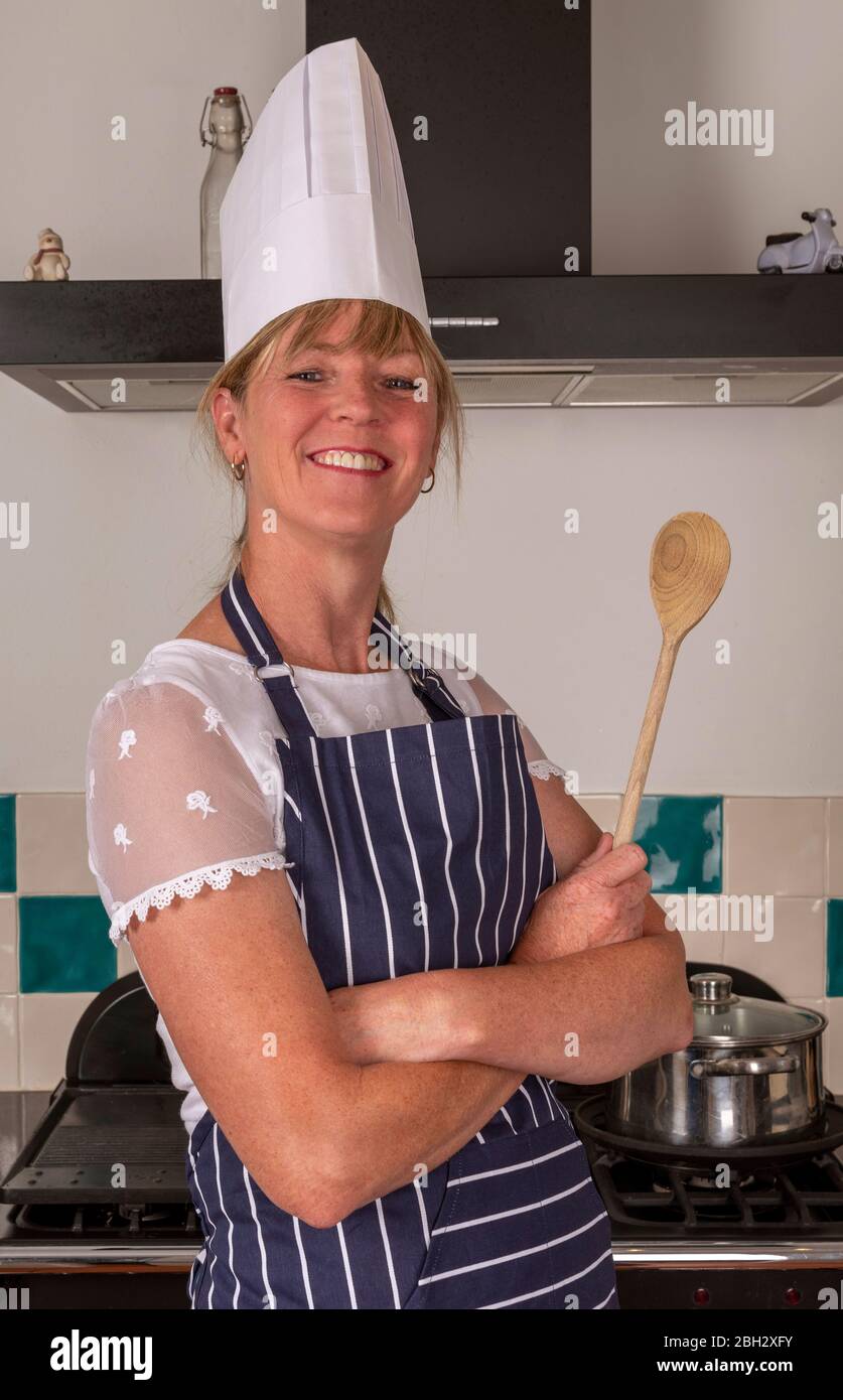 England, Großbritannien. 2019. Attraktive Frau mit einem Holzlöffel und trägt einen Köche weißen Hut Kochen in einer modernen Küche. Stockfoto