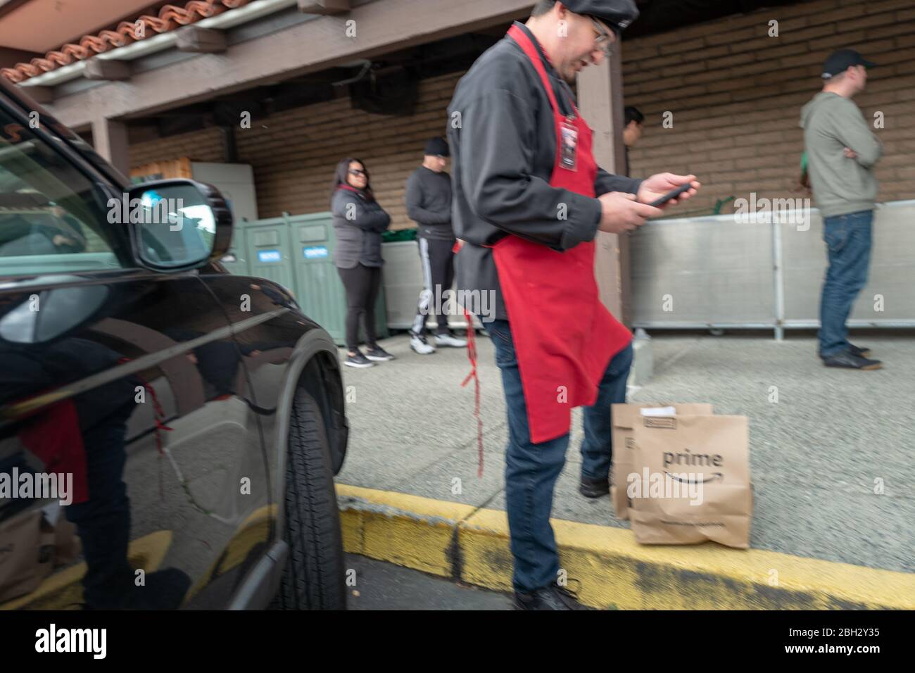 Verschwommenes Motion-Foto eines Mitarbeiters von Whole Foods Market, der einen Amazon Prime Now-Abholservice verarbeitet, während die Leute in der Schlange warten, um einen Whole Foods Market Supermarkt zu betreten, während sie die sozialen Distanzierungsanforderungen während eines Ausbruchs des COVID-19 Coronavirus in Lafayette, Kalifornien, 29. März 2020 beobachten. () Stockfoto