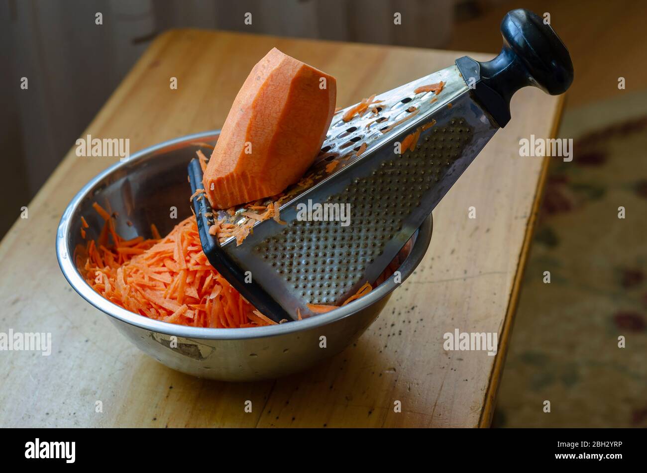 Ein großes Stück Karotten liegt auf einer Reibe. Unterbrochener Zerkleinerungsprozess rohes Gemüse. Der Prozess der Herstellung von Salat. Im echten Leben. Nahaufnahme. Selektiv fo Stockfoto