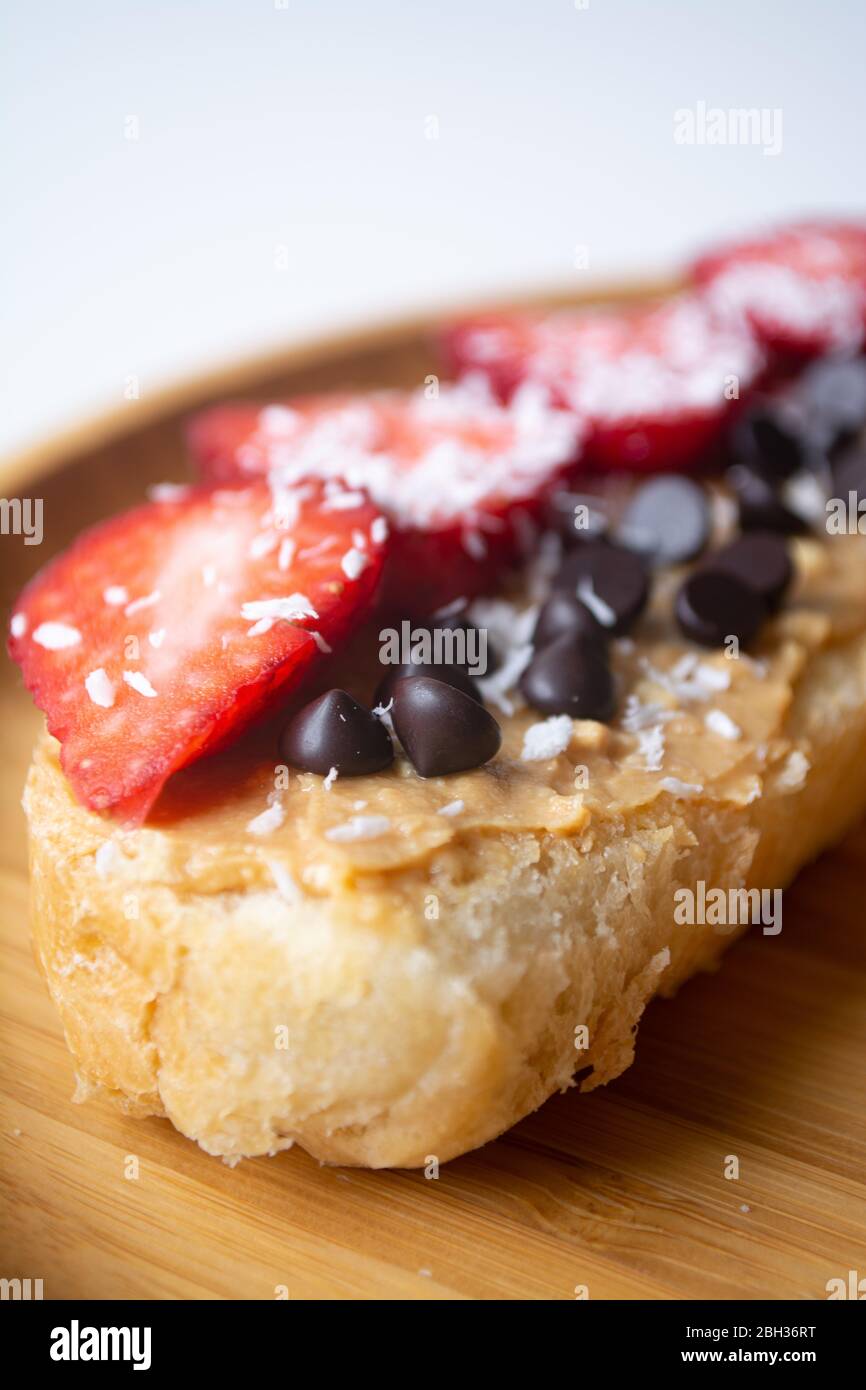 Erdnussbutter Aufstrich mit Schokoladenstückchen, Erdbeeren, Kokosflocken und hausgemachtem Brot auf einem Holzteller Stockfoto