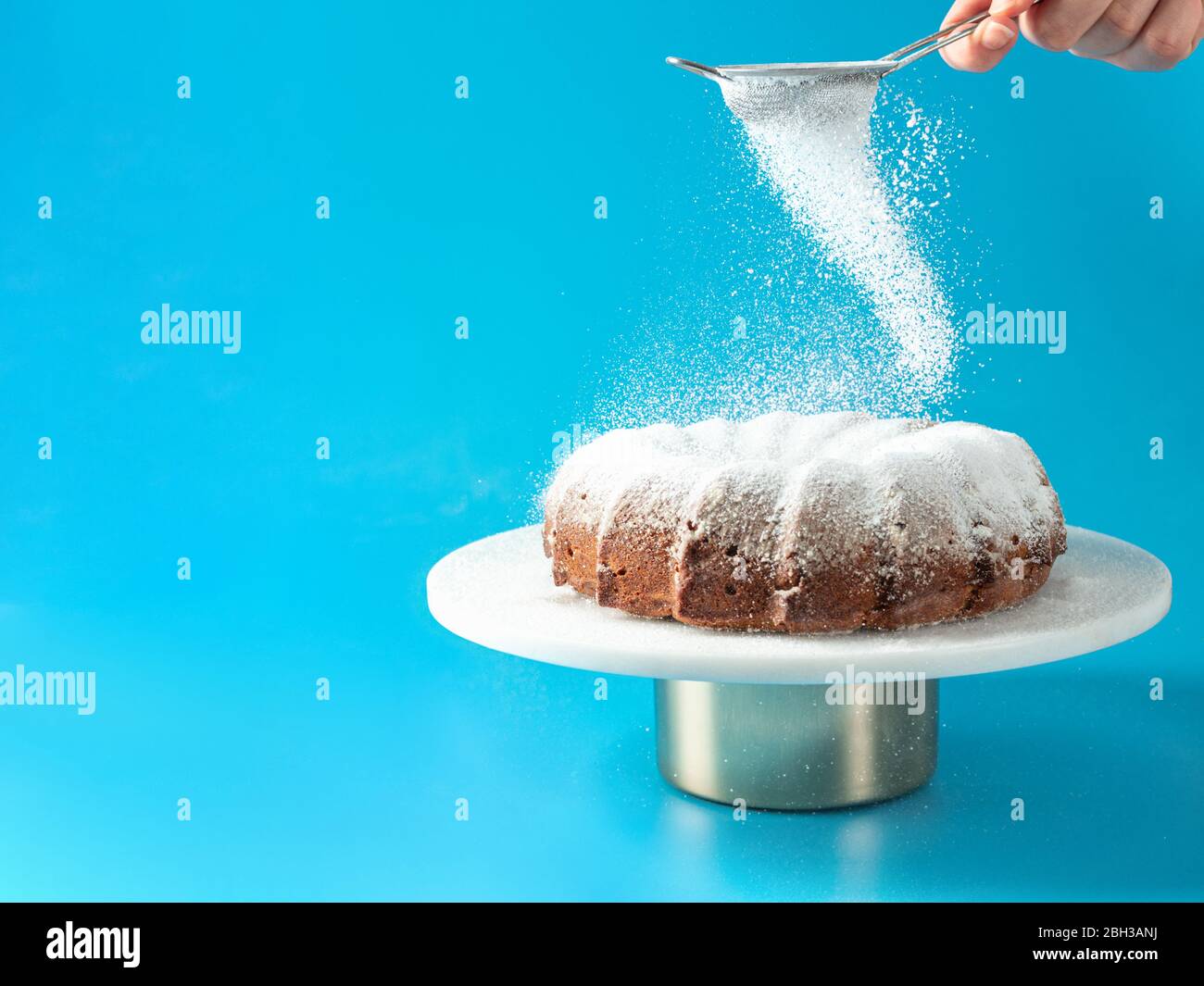 Frau Hand Streuseln Puderzucker über frisch hausgemachte Kuchen gebündelt. Puderzucker fällt auf frischen perfekten Bunt-Kuchen auf blauem Hintergrund. Platz für Text kopieren. Ideen und Rezepte für Frühstück oder Dessert Stockfoto