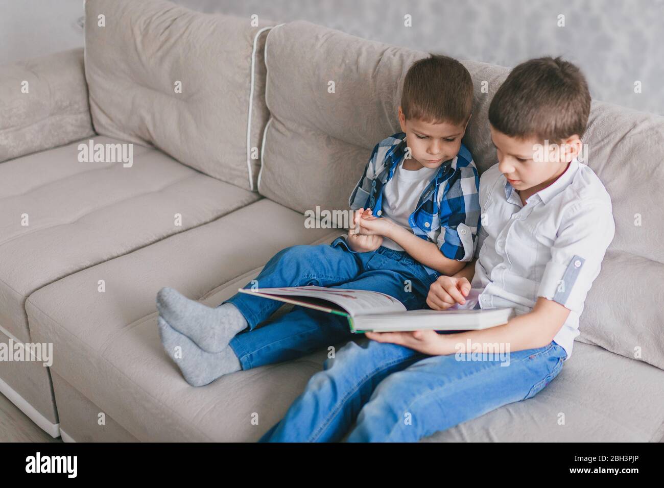 Zwei Brüder lesen ein Buch, während sie während der Quarantäne auf einem Sofa im Wohnzimmer sitzen. Das Konzept der sozialen Distanz, Selbstisolierung und Heimat scho Stockfoto