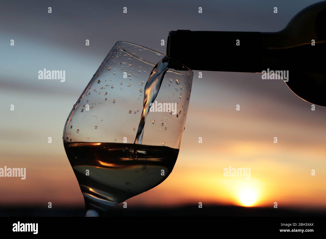 Weißwein, der aus einer Flasche ins Glas strömt auf wunderschönem Sonnenuntergang scheint die orangefarbene Sonne durch die dunklen Wolken. Konzept der Feier Stockfoto