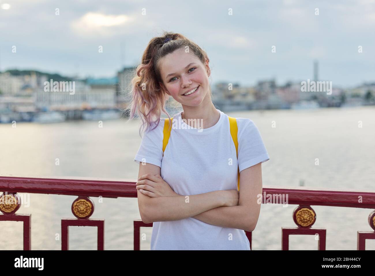 Outdoor-Porträt von lächelnden Teenager-Mädchen von 15, 16 Jahre alt mit gefalteten Armen, Rucksack Blick auf die Kamera. Mädchen auf Brücke stehend, Sonnenuntergang Fluss b Stockfoto