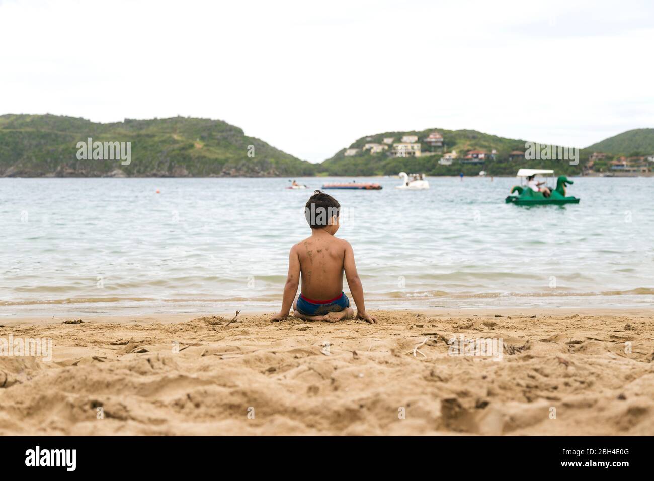Ein einsamer brasilianischer Junge, der auf dem Sand am Strand Brasiliens sitzt. Blick auf seinen Rücken. Strand Von Ferradura, Buzios. Stockfoto
