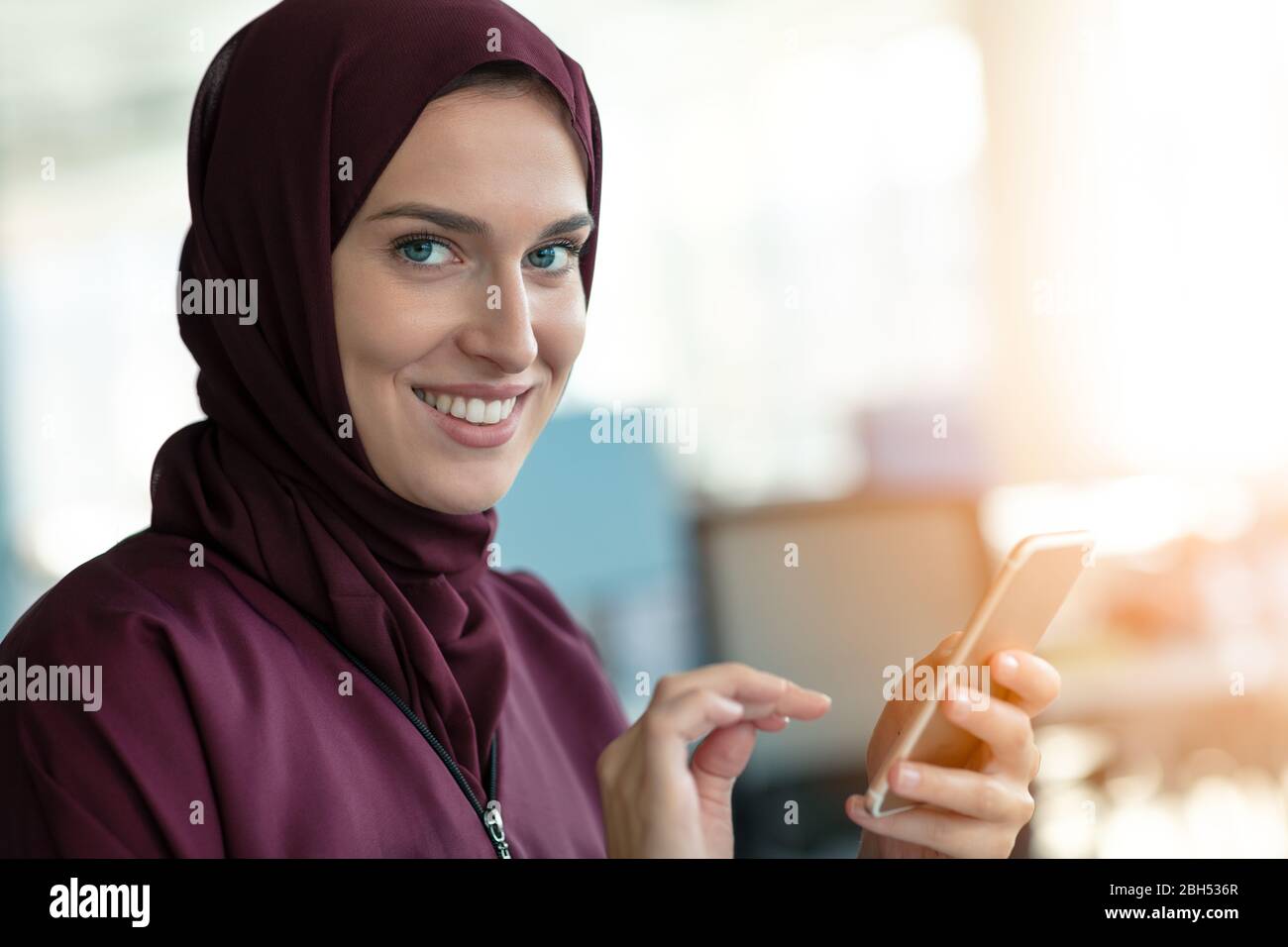 Glückliche muslimische Geschäftsfrau in Hijab am Büroarbeitsplatz. Lächelnde arabische Frau, die am Laptop arbeitet und mit dem Smartphone spricht Stockfoto