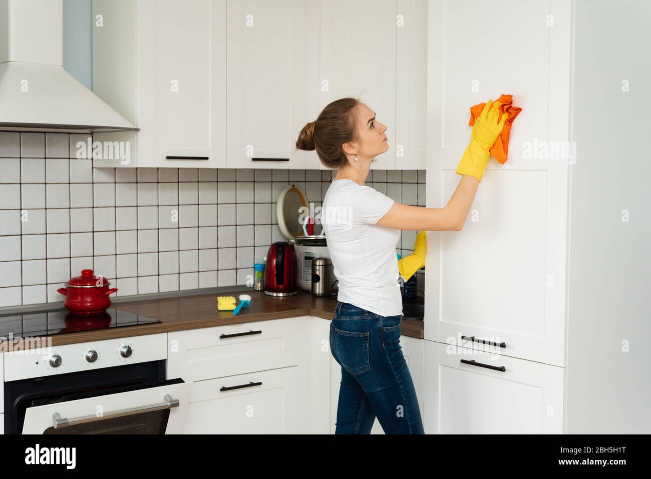 Das Konzept der Frühjahrsputz, Reinigungsfirma. Junge Frau Reinigung einer Oberfläche von weißen Küchenwand Schrank, tragen Gummi-Schutzhandschuhe gelb, mit Lappen. Haus, Hauswirtschaftskonzept. Stockfoto