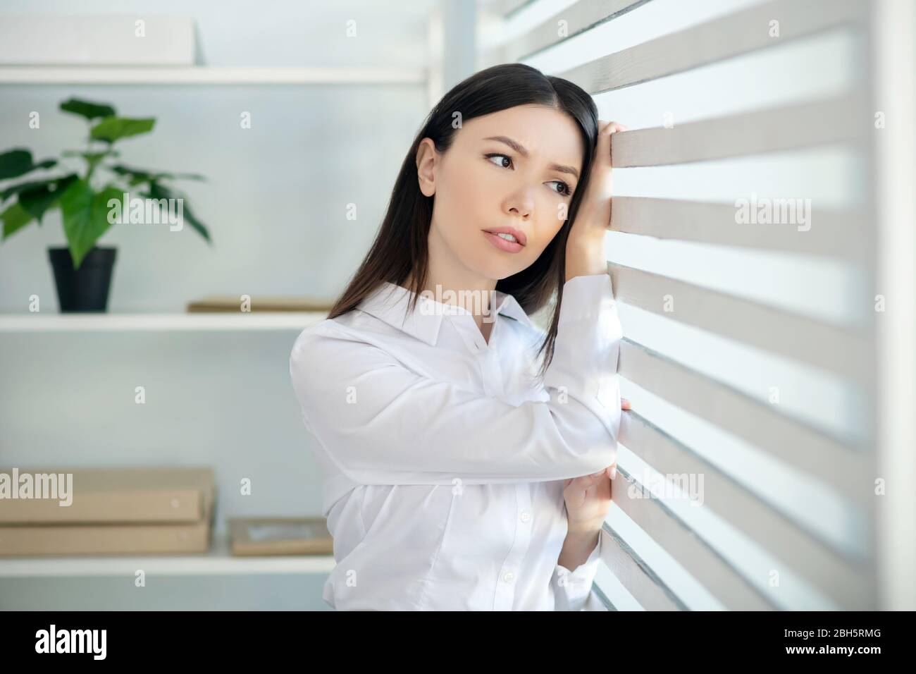 Bei der Arbeit. Junge Brünette Frau steht neben dem Fenster, lehnt ihren Kopf auf Jalousien, berühren sie Stockfoto