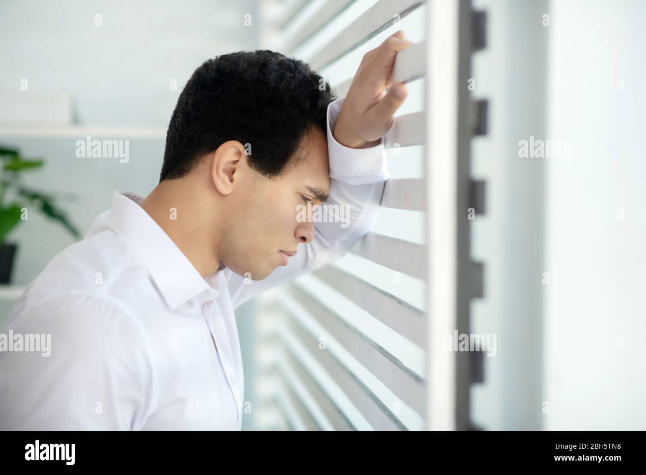 Bei der Arbeit. Verärgert jungen Brünette Mann am Fenster stehen, lehnt seinen Arm und Kopf auf Jalousien Stockfoto