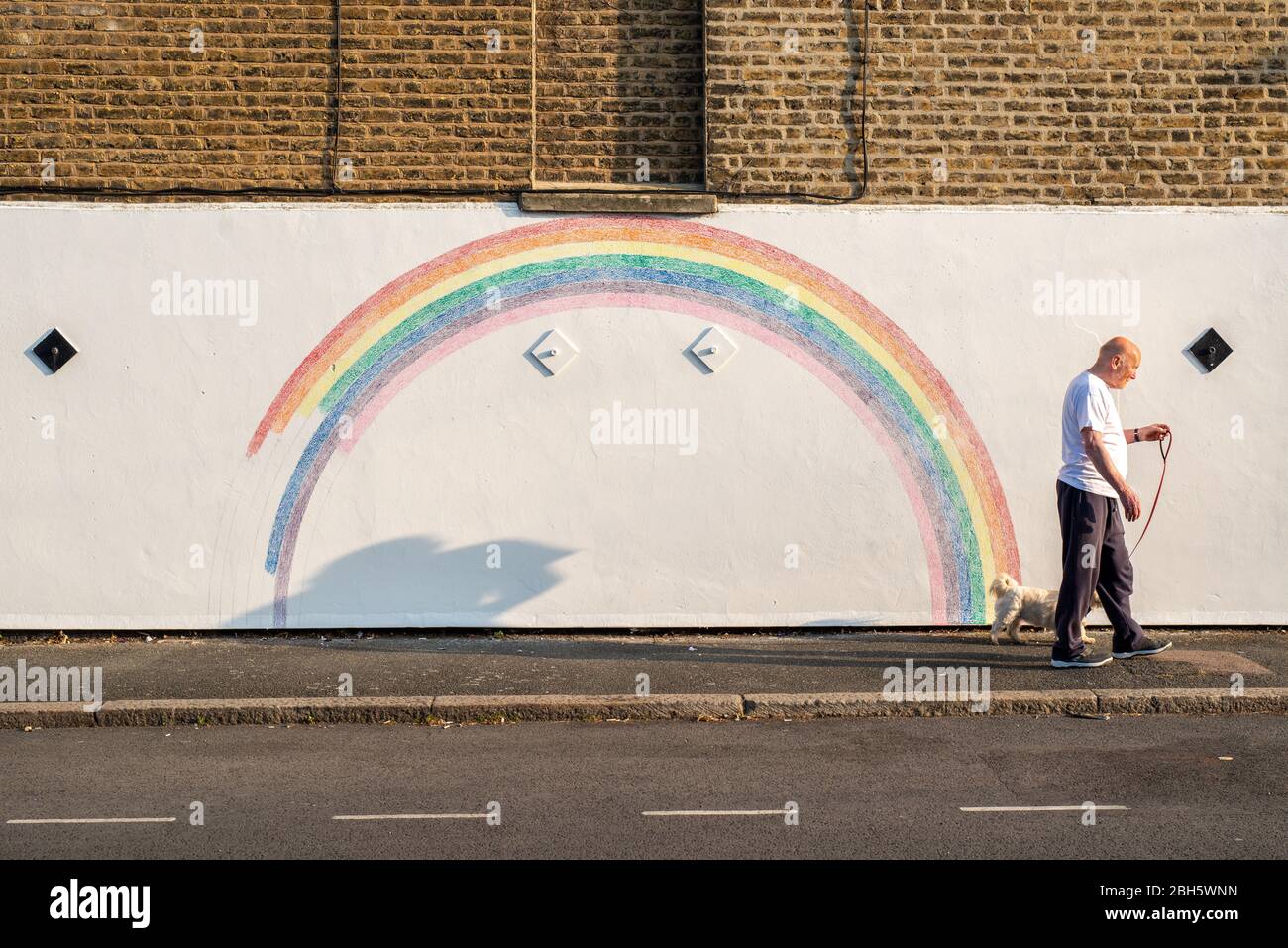 Camberwell, London, Großbritannien. April 2020. Ein Mann geht am frühen Morgen mit seinem Hund an einem Wandbild eines Regenbogens vorbei. Seit der Pandemie Covid-19 ist der Regenbogen zu einem Symbol der Unterstützung für das NHS-Personal und alle Pflegekräfte in Großbritannien geworden. Dieser Regenbogen wird von Louis Young gemalt, um dem NHS-Personal zu danken, das sich um seinen Vater kümmerte und vor kurzem aus dem Krankenhaus kam. Kredit: Tom Leighton/Alamy Live News Stockfoto