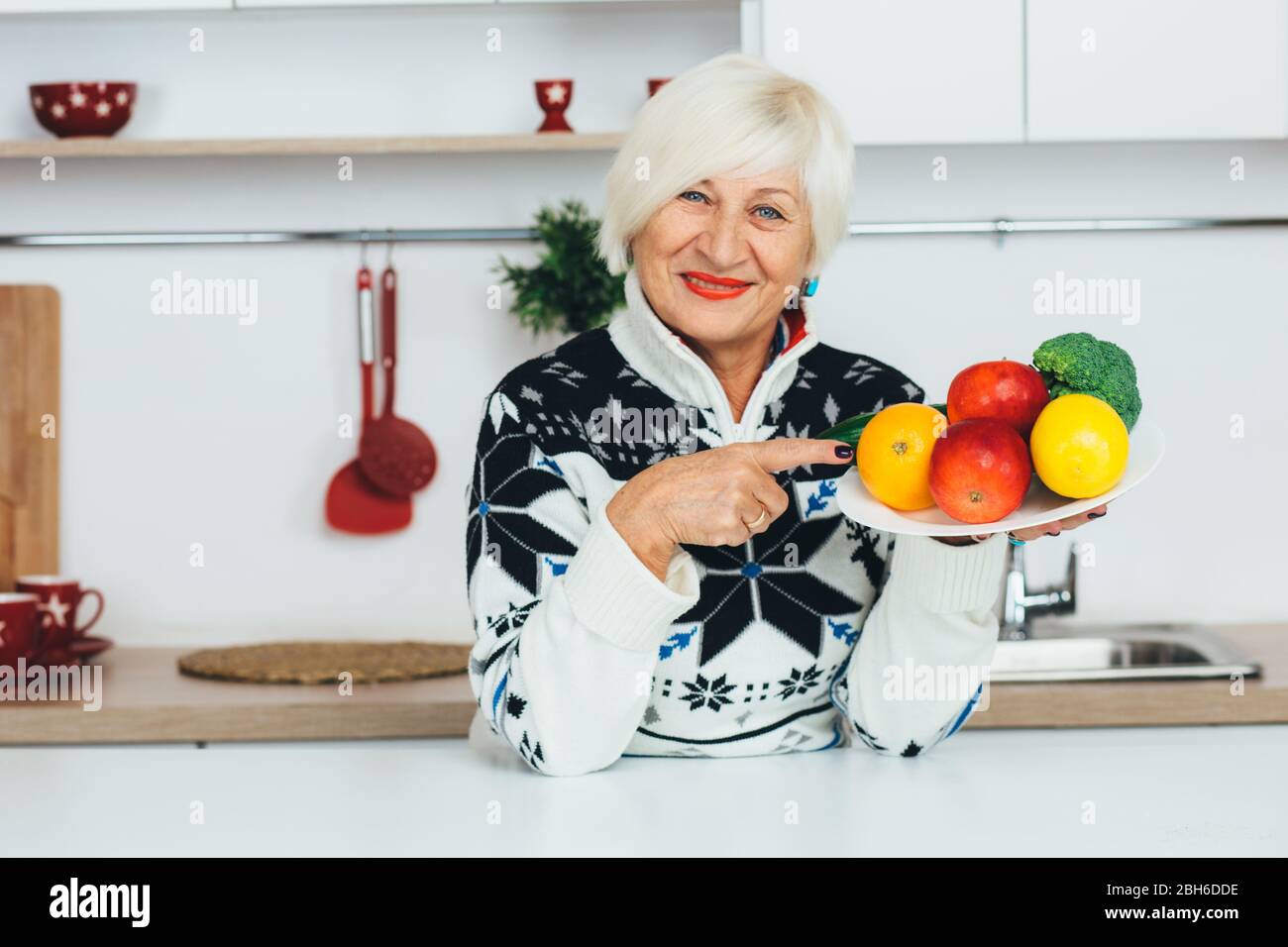 Ältere Frau, die einen Teller mit Obst hält, während sie in der Küche steht und lächelnd die Kamera anschaut. Gesundes Essen für Senioren Stockfoto