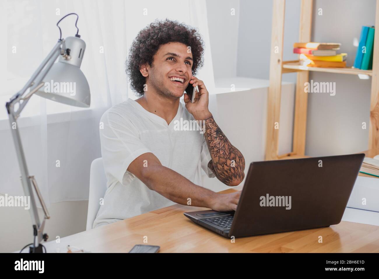 Fröhlicher afrikaner, der am Laptop arbeitet und am Handy spricht, während er an seinem Arbeitsplatz im modernen Büro sitzt Stockfoto