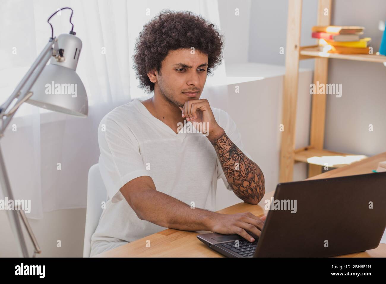 Junger Mann, ein Mulatto mit lockigen Haaren, konzentrierte sich auf Laptop, während er an seinem Arbeitsplatz zu Hause sitzt Stockfoto