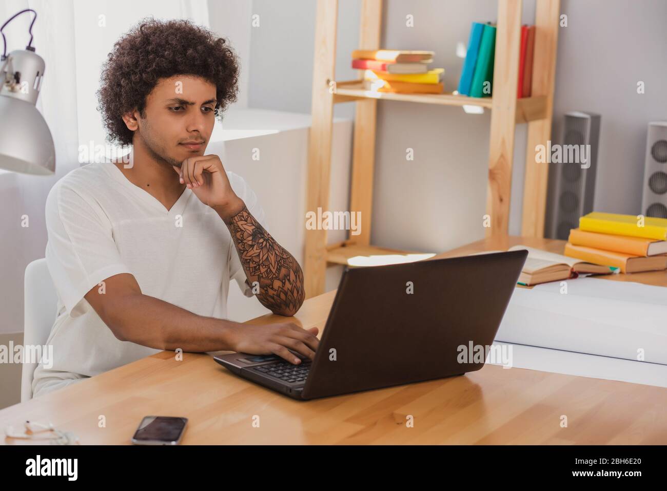 Nachdenklicher Mann, mit lockigen Haaren und gekleidet informell, aufmerksam auf Laptop schauen, während er an seinem Arbeitsplatz zu Hause sitzt Stockfoto