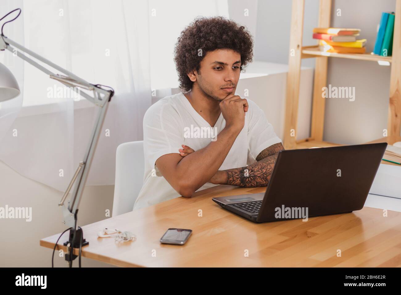 Nachdenklicher Mann, mit lockigen Haaren und gekleidet informell, aufmerksam auf Laptop schauen, während er an seinem Arbeitsplatz im Büro sitzt Stockfoto