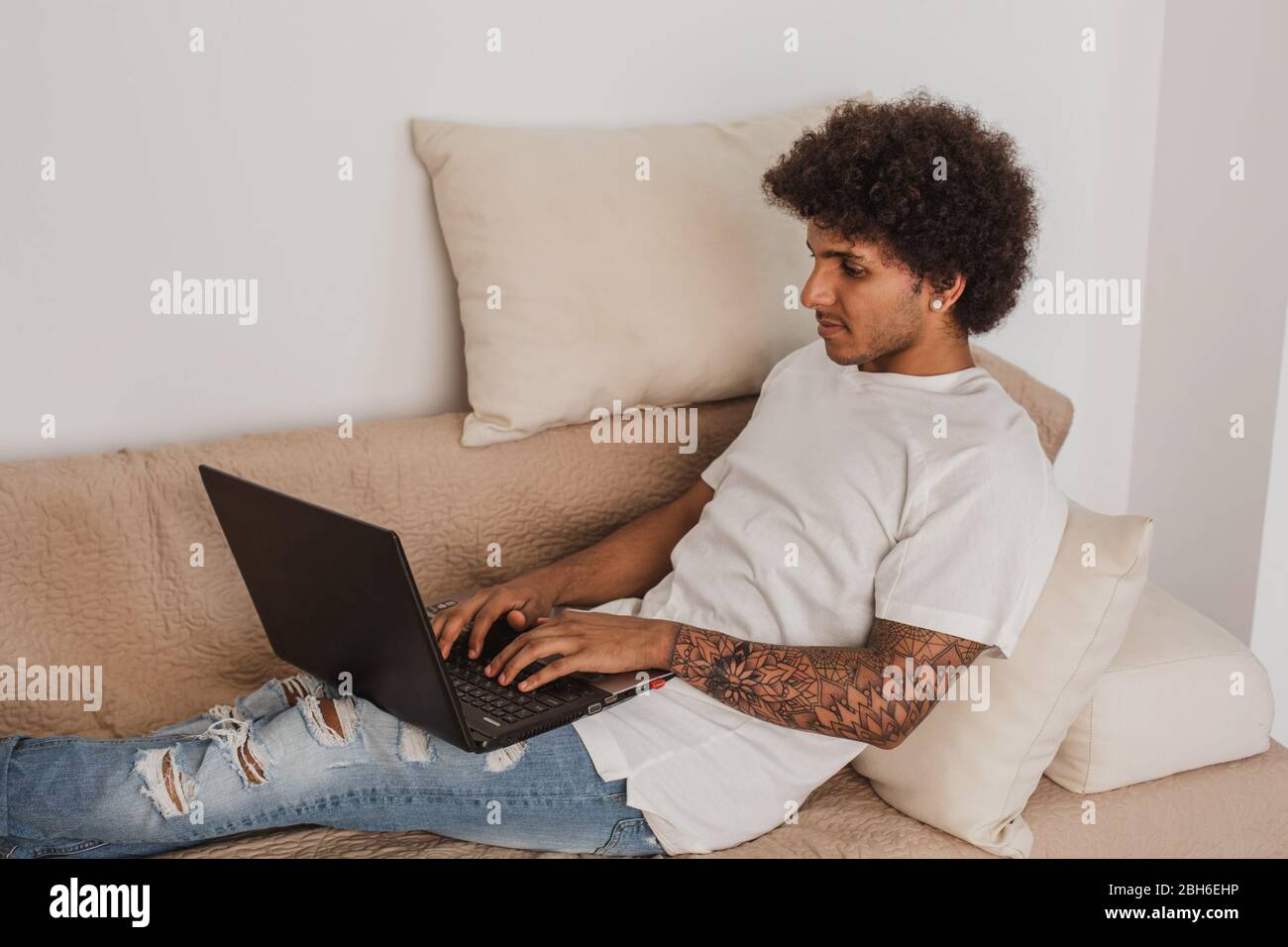 Junger Mann, ein Mulatto mit lockigen Haaren, entspannt auf der Couch mit einem Laptop, zu Hause Stockfoto