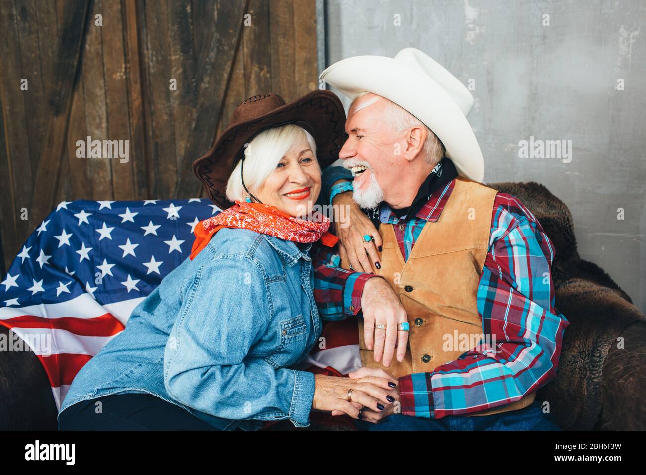Ältere Paare, in Cowboyhüte gekleidet, lachen, nehmen sich die Hände. Hinter, auf der Couch liegt die amerikanische Flagge, die Feier von Amerika in Stockfoto