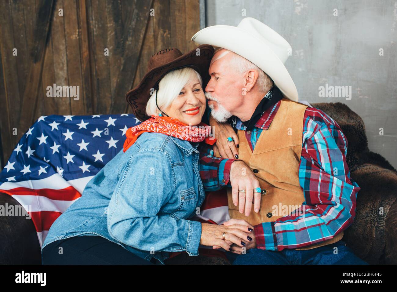 Ältere Paare, in Cowboyhüte gekleidet, lächelnd, sich gegenseitig die Hände nehmend. Hinter, auf der Couch liegt die amerikanische Flagge, die Feier der amerikanischen Stockfoto