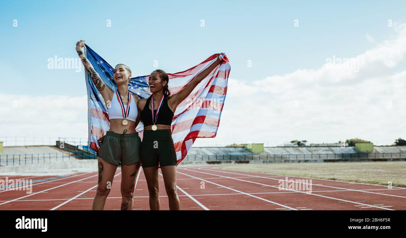 Zwei Athletinnen feiern den Sieg mit amerikanischer Flagge auf der Laufstrecke. Glückliche Frauen Athlet schreiend vor Freude hält die US-Flagge hinter ihnen A Stockfoto