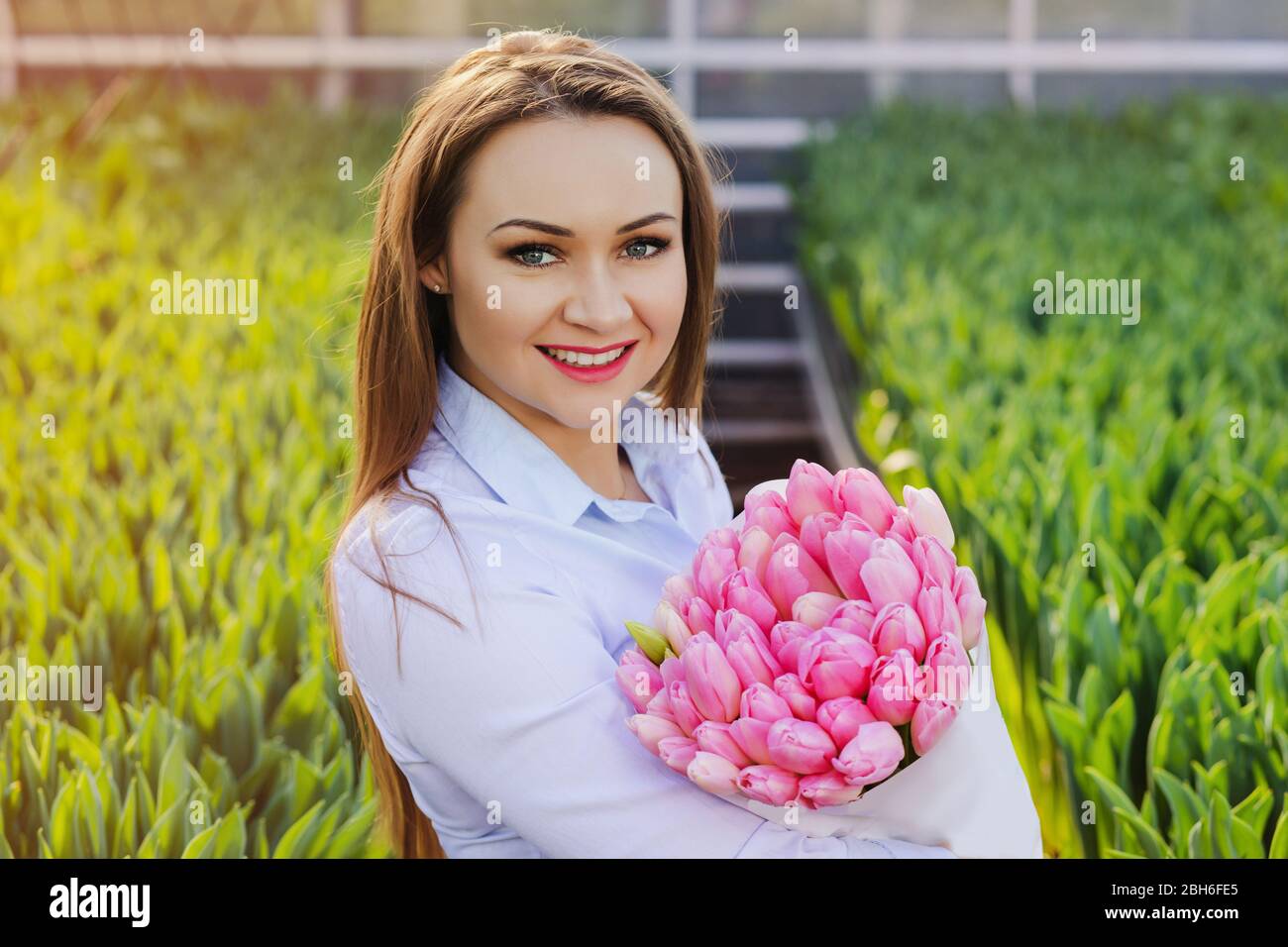 Schöne junge Frau mit einem perfekten Lächeln, hält einen großen Strauß Tulpen, Blick auf die Kamera, in einem Gewächshaus stehen Stockfoto