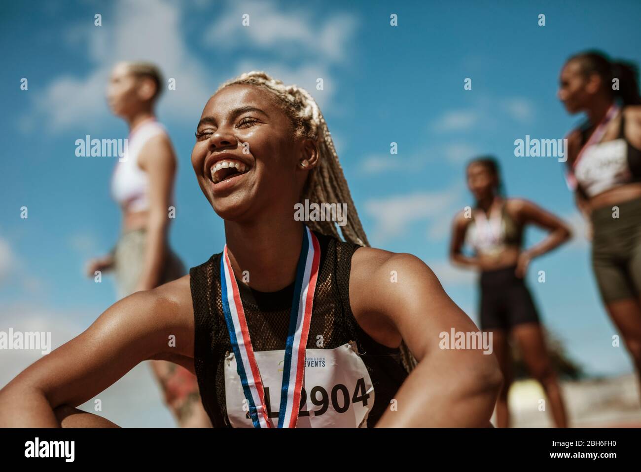 Athletinnen lächeln, nachdem sie ein Rennen mit anderen Konkurrenten im Hintergrund gewonnen haben. Sportlerin mit Medaille zum Sieg im Stadion. Stockfoto