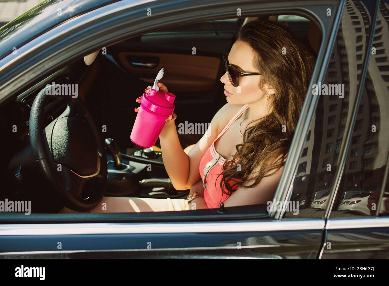 Erfolgreiche Frau, in lässiger Kleidung gekleidet, trinkt Sportnahrung, sitzt in einem luxuriösen Auto. Gesunde Ernährung überall Stockfoto